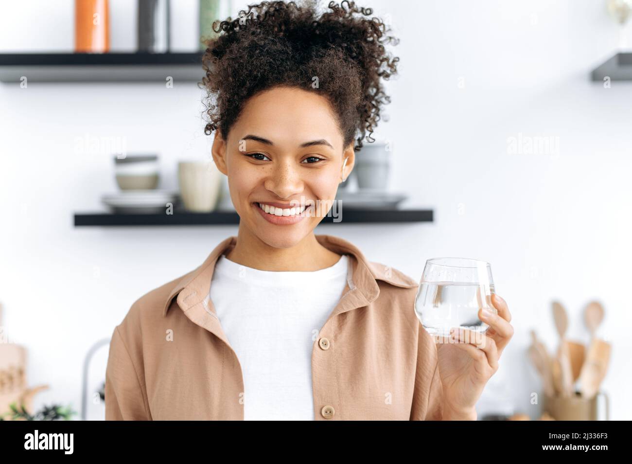 Glückliches gesundes afroamerikanisches Mädchen in Freizeitkleidung, zu Hause in der Küche stehend, hält ein Glas sauberes Wasser, trinkt täglich eine Menge Wasser, kümmert sich um ihre Gesundheit, schaut auf die Kamera, lächelt Stockfoto