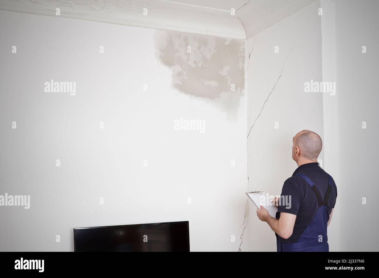 Ein Hausmeister oder Hausmeister inspiziert einen großen Wasserfleck und verschiedene Risse in Wand und Decke des Wohnzimmers eines alten Hauses. Stockfoto