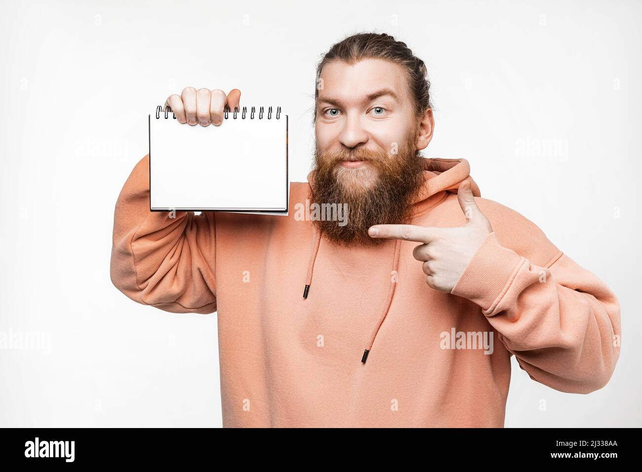 Skandinavischer gutaussehender glücklicher Mann hält Notizblock und zeigt auf Papiermockup isoliert auf grauem Hintergrund. Kerl mit Ingwer-Frisur und Bart im Schweiß Stockfoto