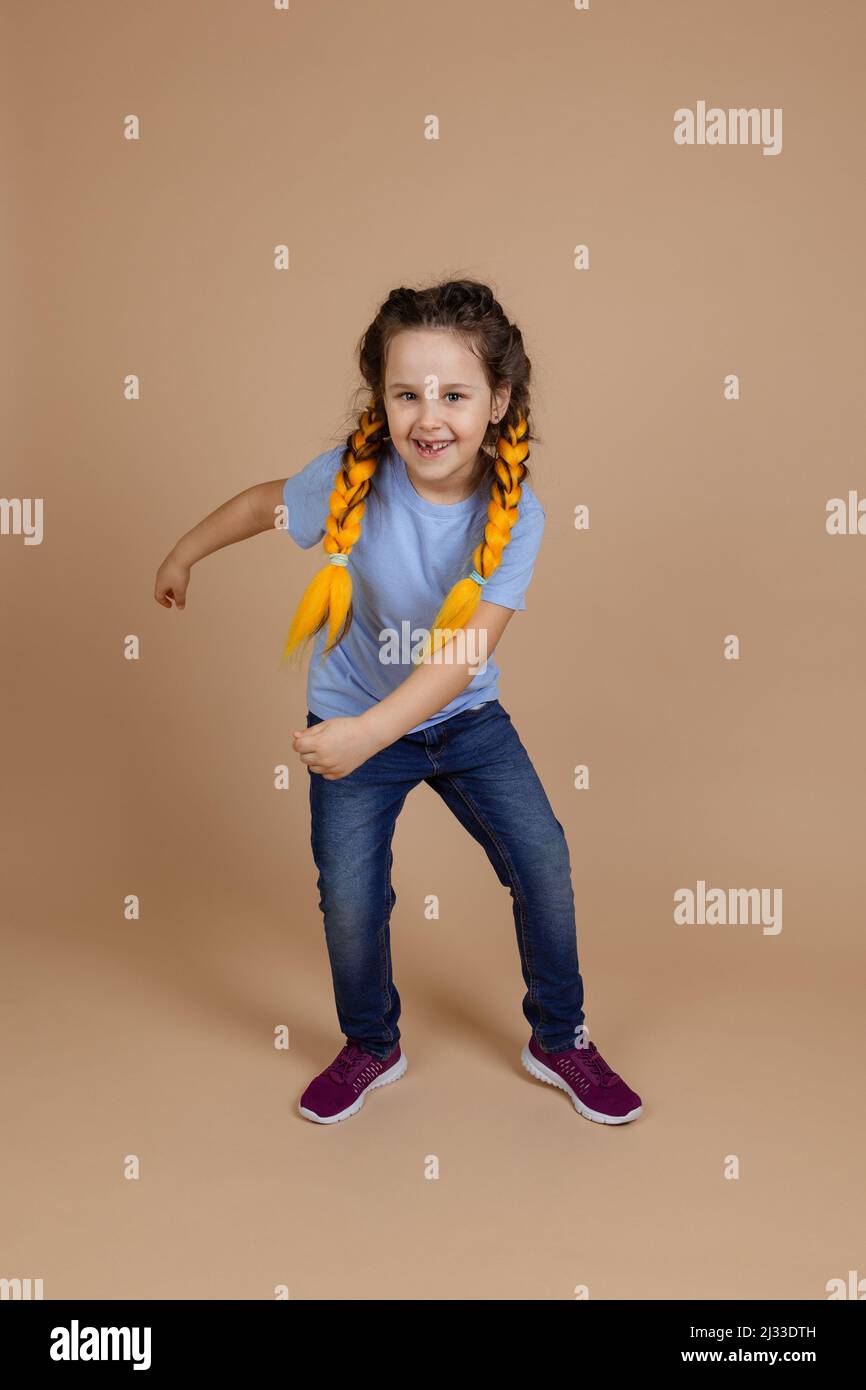 Leuchtendes kaukasisches kleines Mädchen mit gelben Kanekalon-Zöpfen, tanzenden beweglichen Armen, lächelnd mit fehlendem Zahn und Blick auf die Kamera auf beigefarbenem Hintergrund Stockfoto