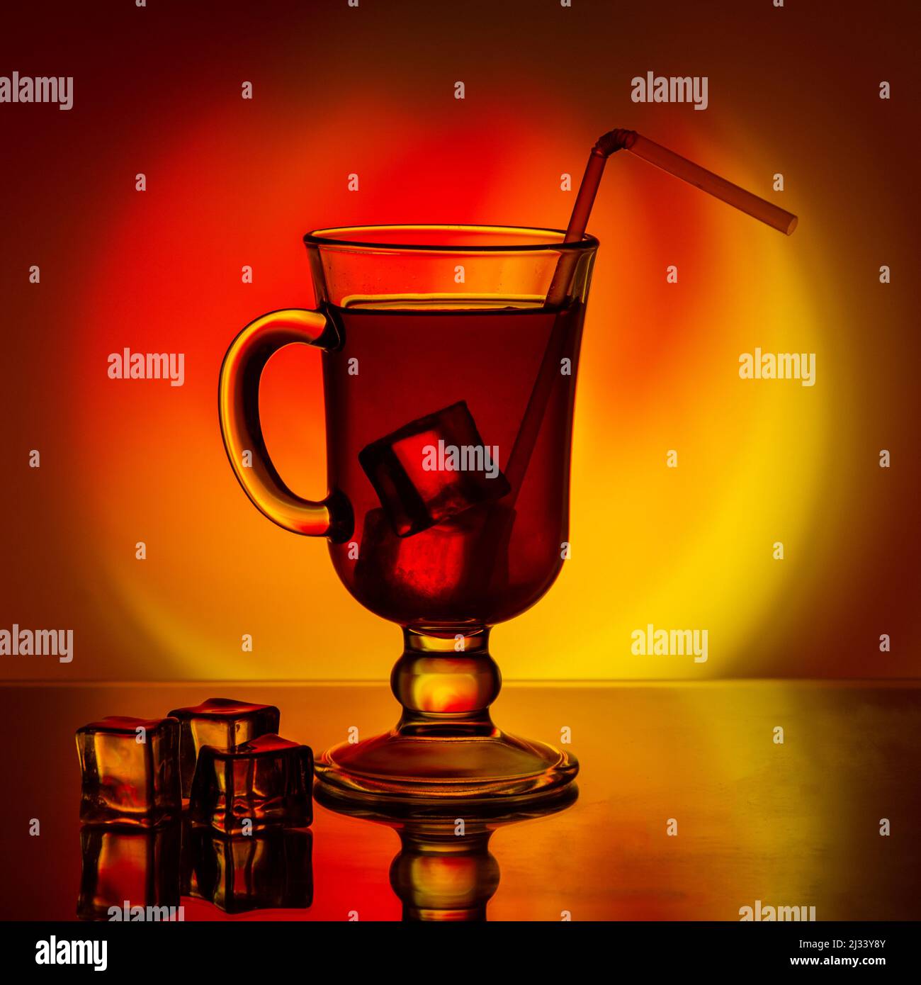 Kalter Tee, Cocktail, Fruchtgetränk in einer hohen Glastasse mit Trinkhalm auf einem Tisch mit Eisstücken auf einem schönen, leuchtend gelb-roten Farbverlauf zurück Stockfoto