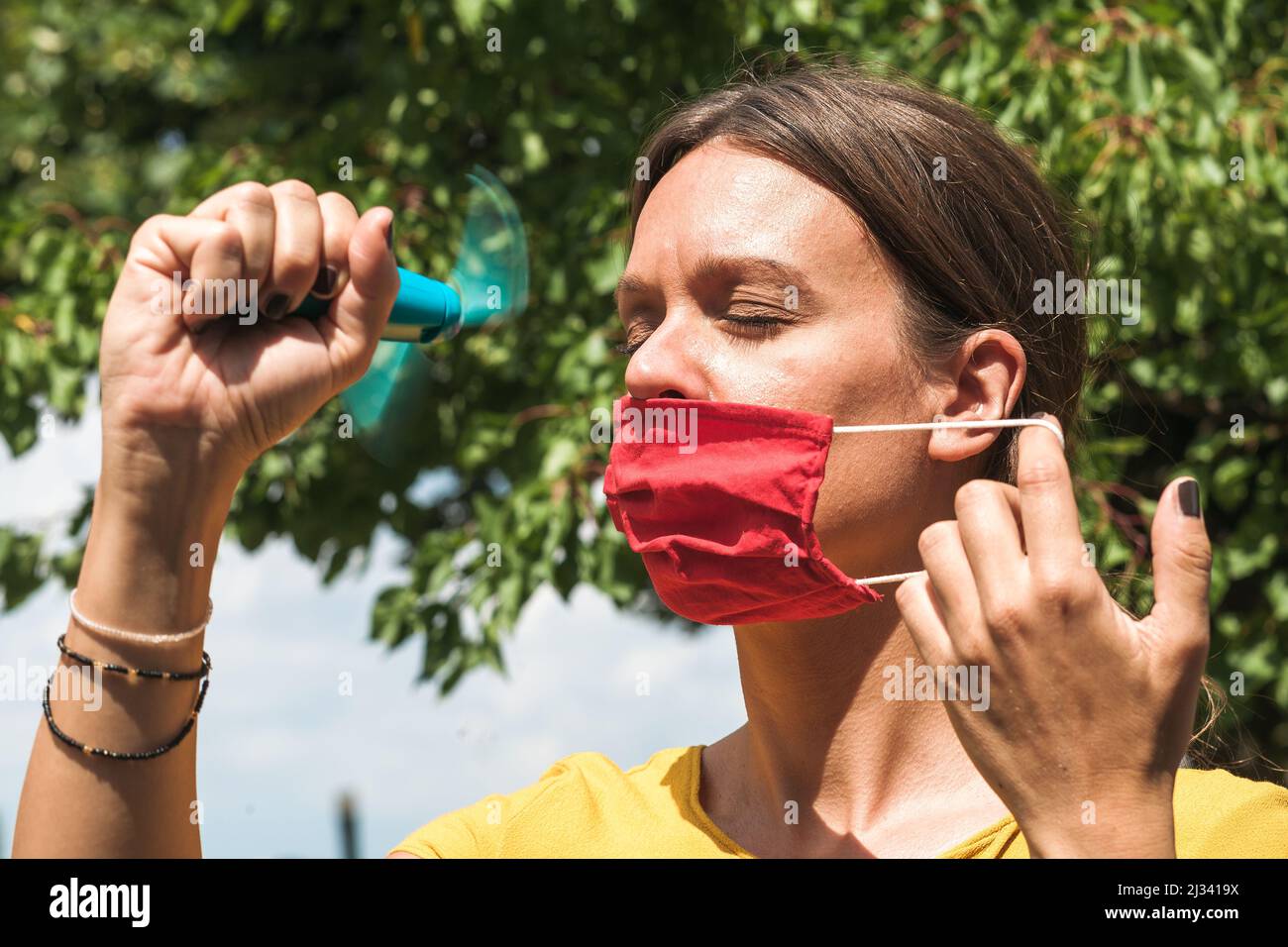 Junge Frau kühlt sich mit einem Handventilator ab, während sie eine Gesichtsschutzmaske trägt Stockfoto