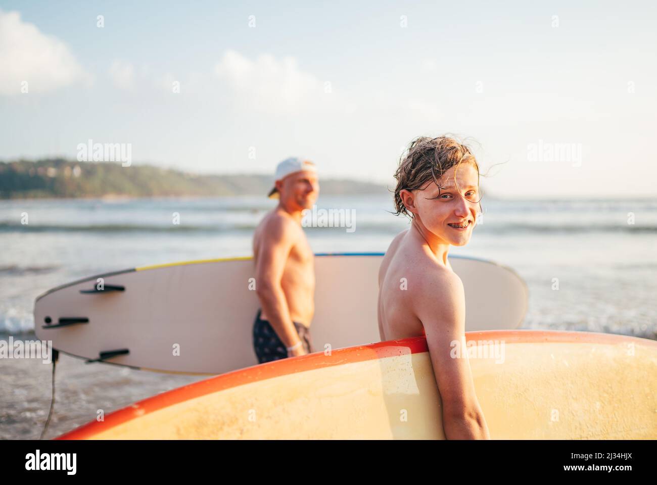 Lächelnder Teenager-Junge mit Surfbrett geht zum Surfen ins Meer. Er hat einen Winterurlaub und genießt ein schönes Sonnenuntergangslicht mit Vater auf Sri Lanka ist Stockfoto