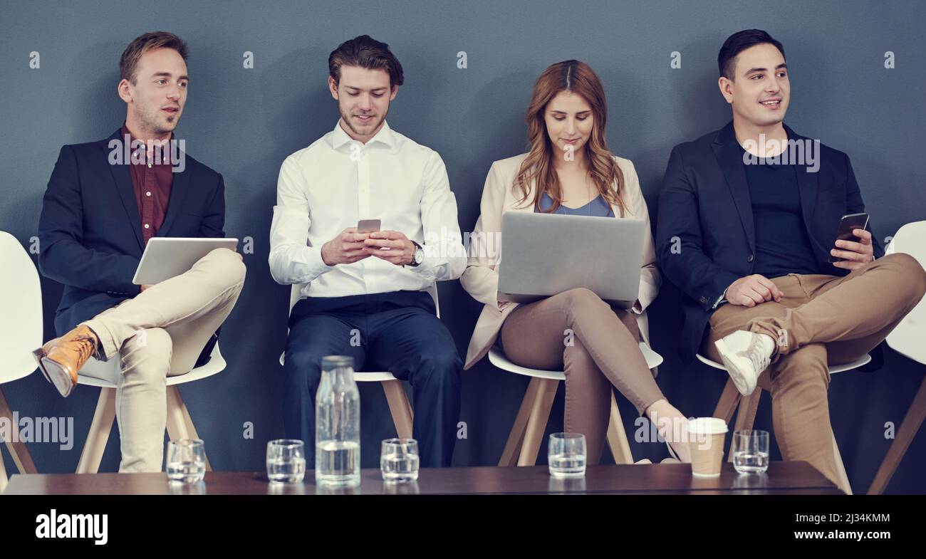 Auf ihren drahtlosen Geräten während des Wartens. Aufnahme einer Gruppe von Geschäftsleuten, die verschiedene drahtlose Geräte verwenden, während sie in der Schlange auf ein Interview warten. Stockfoto