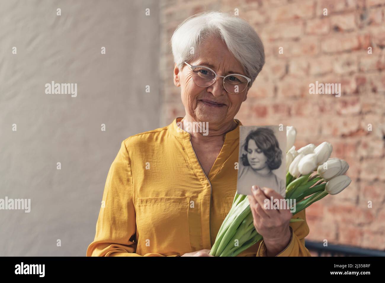 Oma's Day – mittlere Aufnahme einer Großmutter in einem sonnigen gelben Hemd mit Blumen und einem schwarz-weißen Foto ihrer jüngeren Version. Hochwertige Fotos Stockfoto