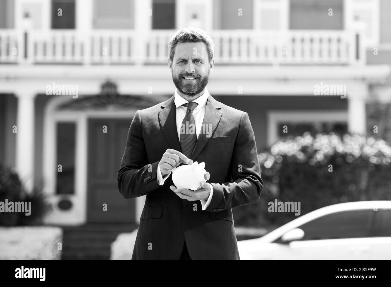 Glücklicher junger Mann, der Geld in ein Sparschwein gesteckt hat. Geschäftsmann hält Sparschwein auf reichem Haushintergrund. Sparen Sie Geld und finanzielle Investitionen. Stockfoto
