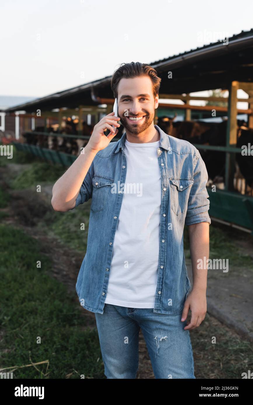 Bauer spricht auf dem Smartphone in der Nähe von verschwommenem Cowhouse und lächelt der Kamera zu Stockfoto