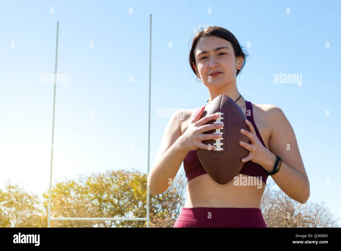 Eine junge kaukasische Athletin, die einen amerikanischen Fußballball an den Seilen hält. Sie trägt ein Aktivitätsarmband und in Th sind Torpfosten sichtbar Stockfoto
