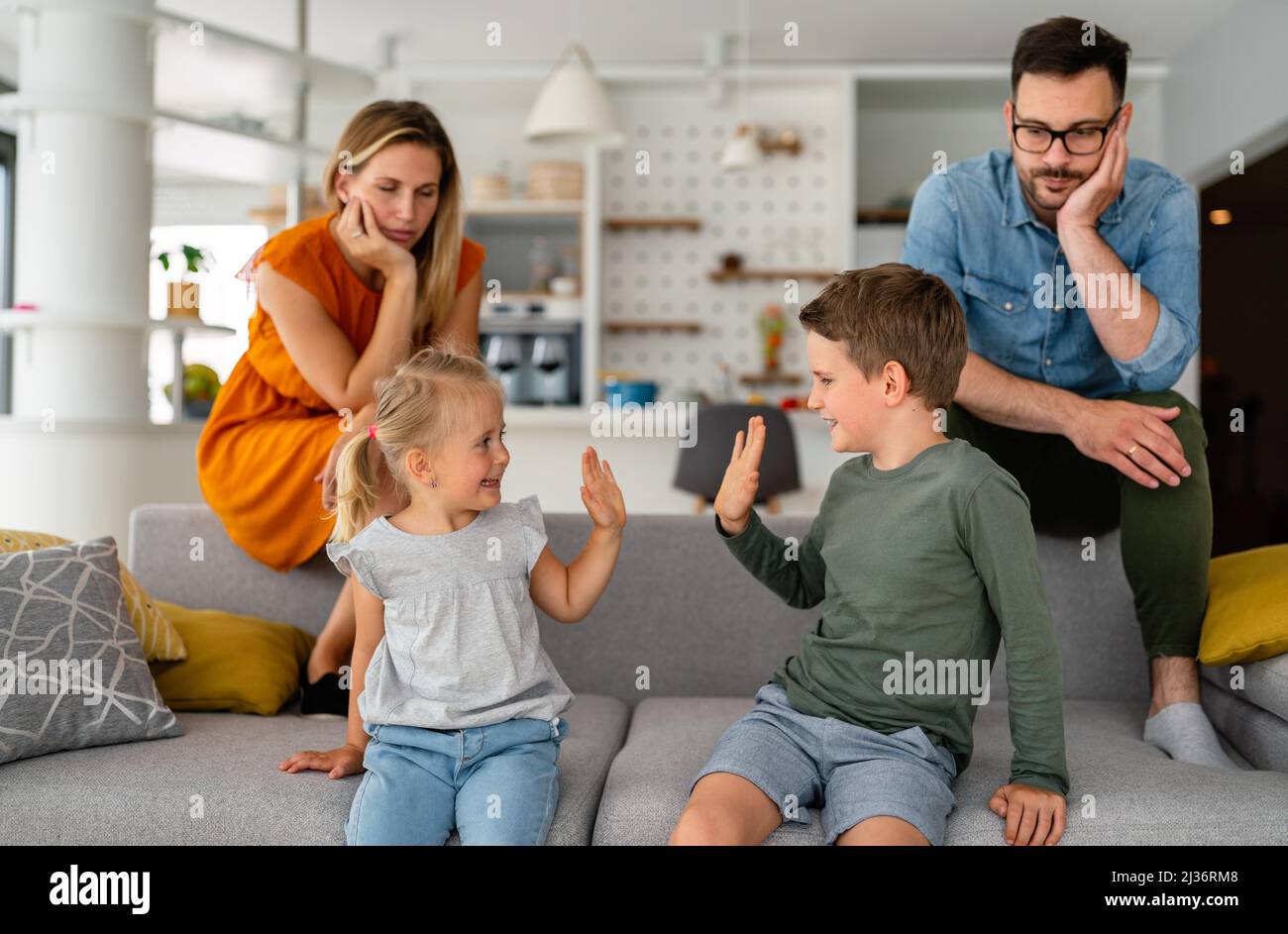 Müde Eltern, die auf der Couch sitzen, fühlen sich erschöpft genervt, während glückliche Kinder zusammen spielen. Stockfoto
