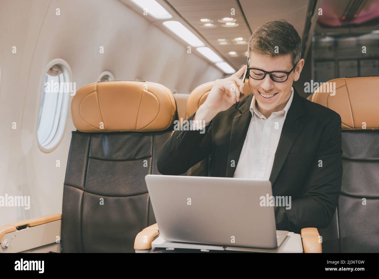 Geschäftsmann, der auf dem Laptop über Satelliten-Internet telefoniert und während der Flugreise einen Geschäftskontakt anruft, hat ein glückliches Lächeln auf den Lippen. Stockfoto