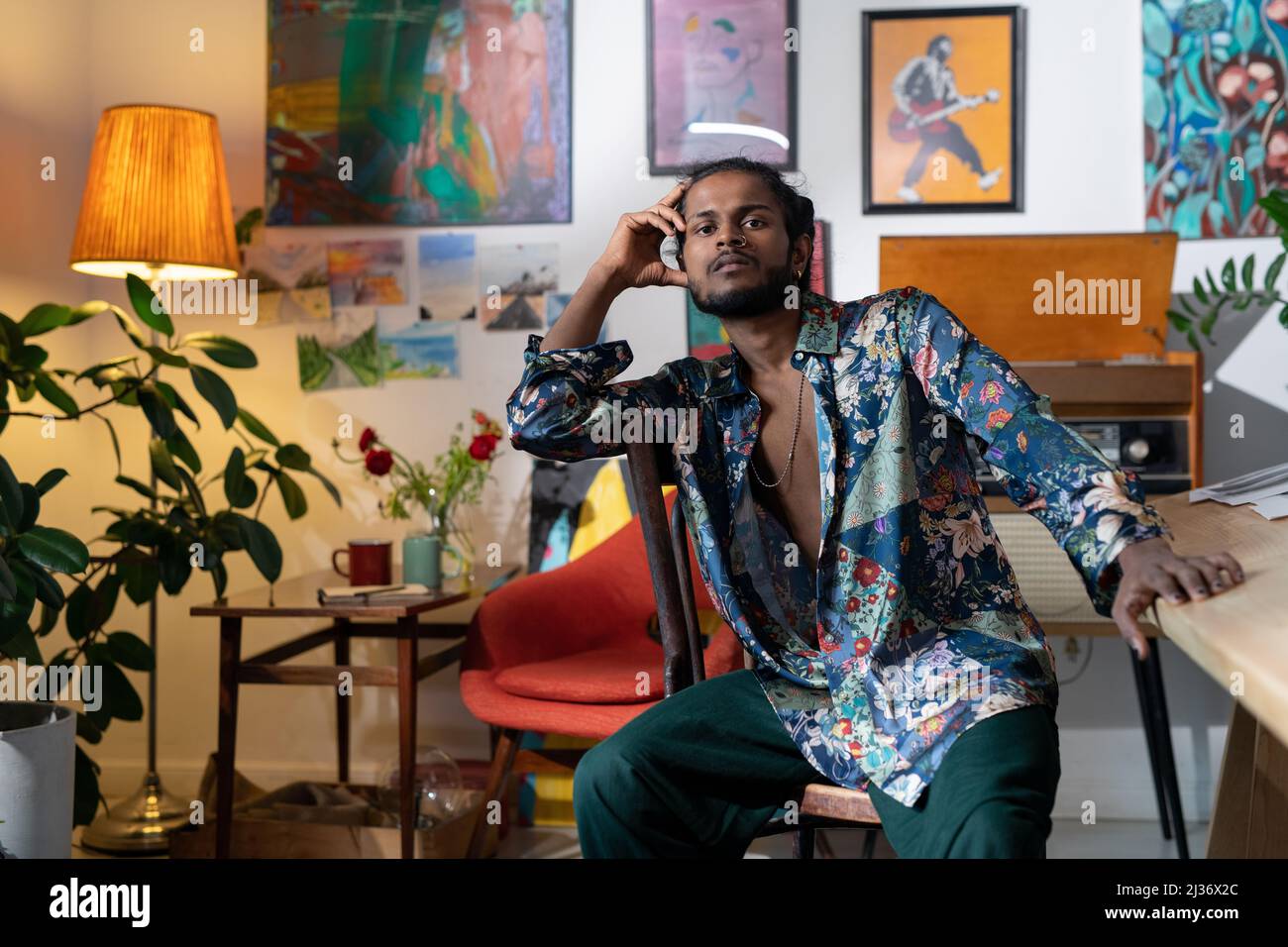 Porträt eines hübschen jungen indischen Mannes mit buntem Hemd, der im Wohnzimmer sitzt, das mit Gemälden geschmückt ist Stockfoto
