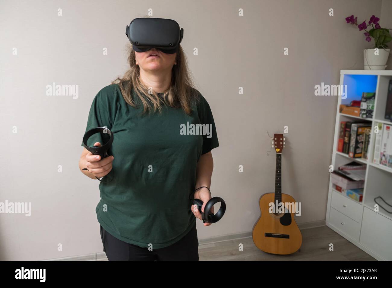 Ein Mädchen in einem grünen T-Shirt mit VR-Brille und Controllern in ihren Händen reist durch das Metaglas-Universum, während es in einem grauen Raum steht. Ein Virt Stockfoto