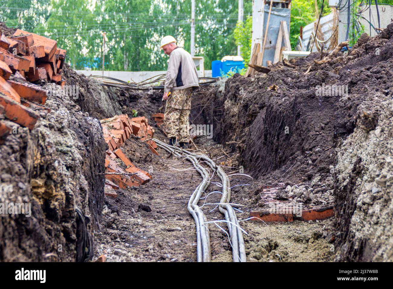 Kemerowo, Russland - 30. juni 2021. Bauingenieur oder Vorarbeiter in einem weißen Schutzhelm steuert ein elektrisches Stromkabel, das in einem Graben verlegt ist, selektiver FOC Stockfoto
