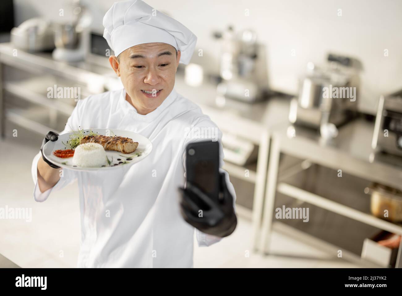 Der asiatische Koch zeichnete am Telefon einen kulinarischen Vlog auf und zeigte einen Teller mit gekochtem Essen in der Küche. Konzept von Video-Blogging und Kochen von Mahlzeiten für das Restaurant Stockfoto