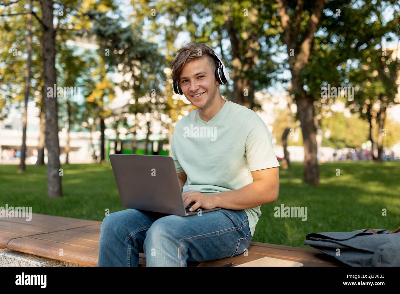 Aufgeregter kaukasischer Kerl, der sich mit einem Laptop und Kopfhörern im Park entspannt, auf einer Bank sitzt und im Freien lächelt Stockfoto