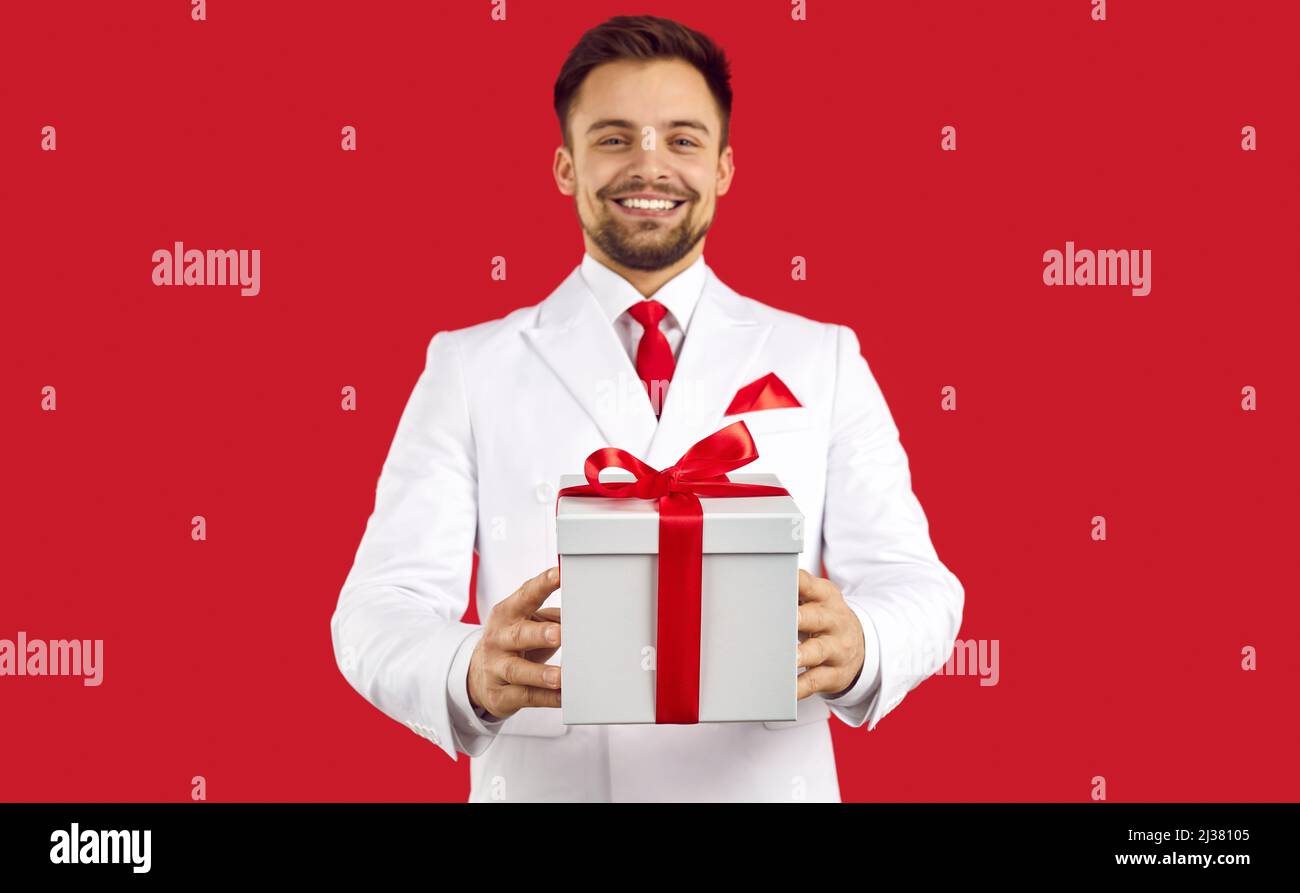 Ein hübscher Mann in einem Anzug, der auf einem roten Studiohintergrund steht und eine weiße Präsens-Box hält Stockfoto