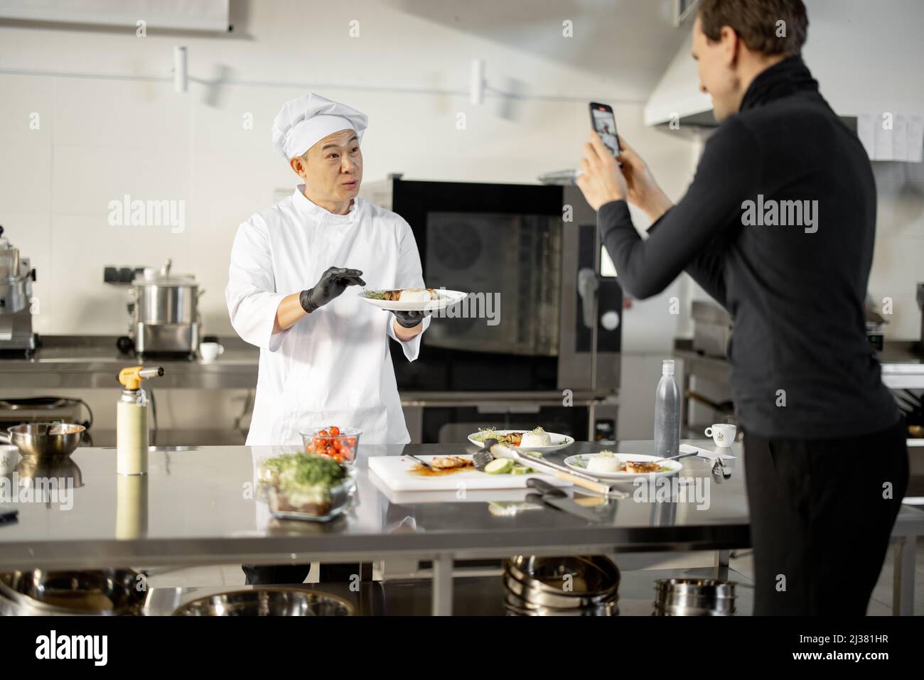 Asiatischer Koch in Uniform spricht über das gekochte Gericht, während er einen kulinarischen Blog in der professionellen Küche unterhält. Mann zeichnet ihn am Telefon auf. Konzept des kulinarischen Videobloggens Stockfoto