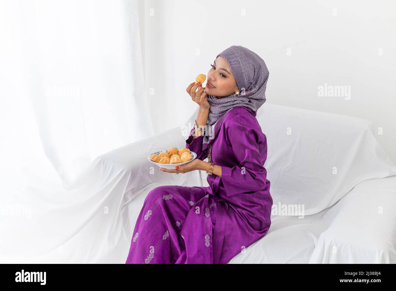 Eine junge malaiische Dame mit Hijab-Kopfbedeckung feiert das Ende des Ramadan, bekleidet mit purpurpurem Kebaya-Kleid, mit traditionellem Bahulu-Kuchen, die Hüfte nach oben Stockfoto