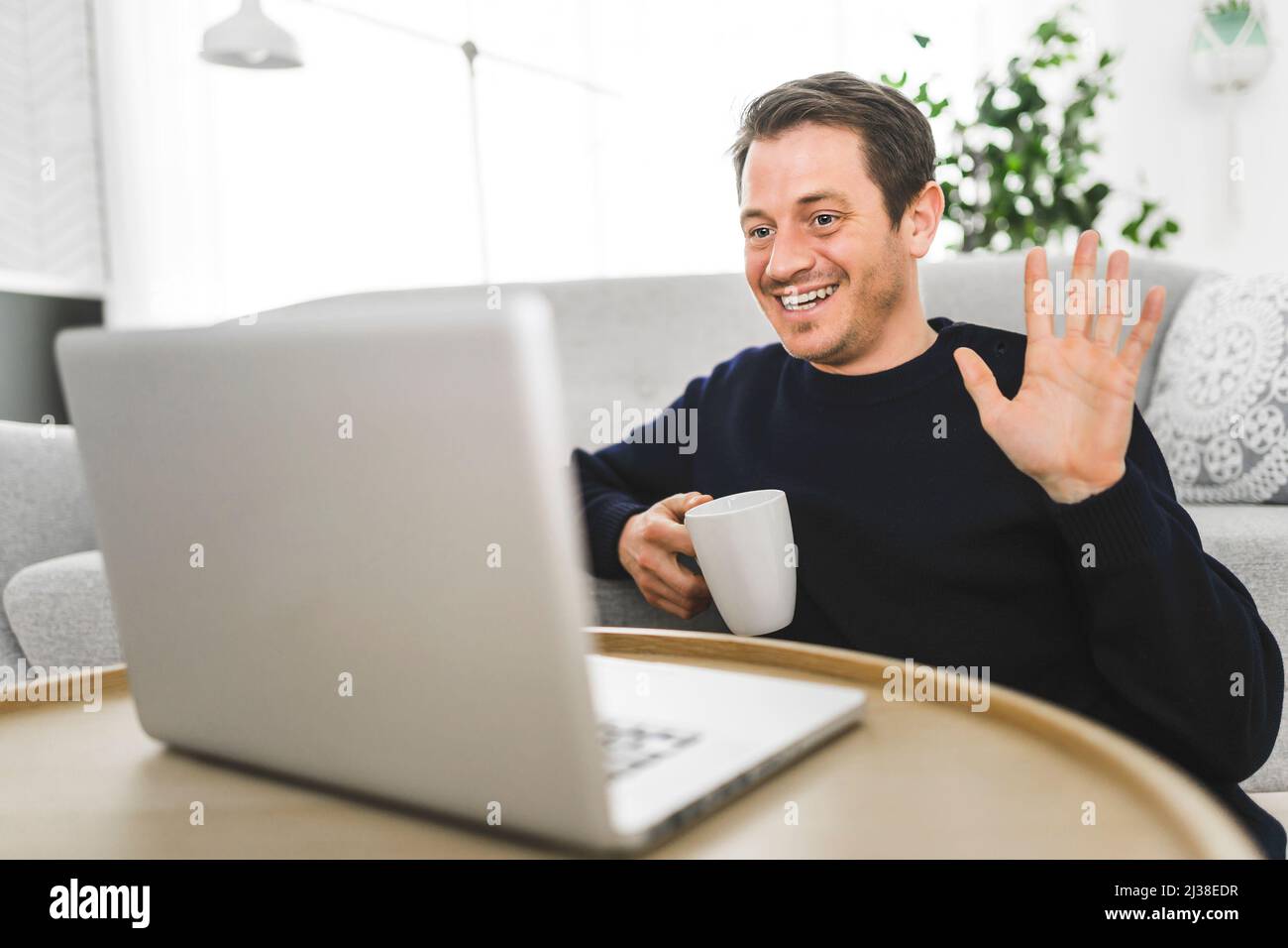 Europäischer Mann, der im hellen Wohnzimmer auf seinem Laptop Video-Chat macht, sagt Hallo Stockfoto