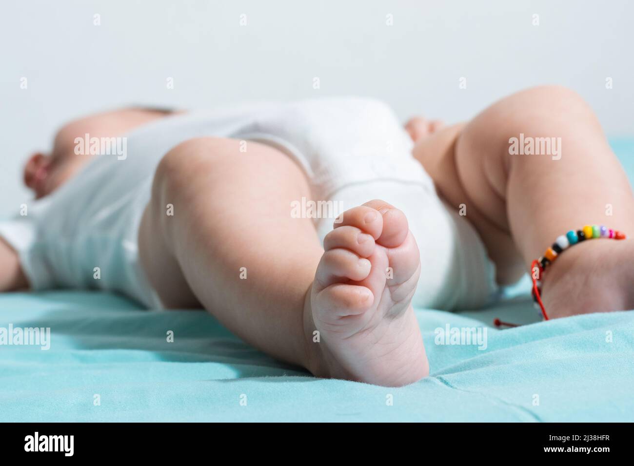Schöne und zarte Füße eines zwei Monate alten Mädchens, mit ihren kleinen Zehen fest eingefahren, ist sie in einem weißen Strampelanzug gekleidet. Auf einem Bett liegend Stockfoto