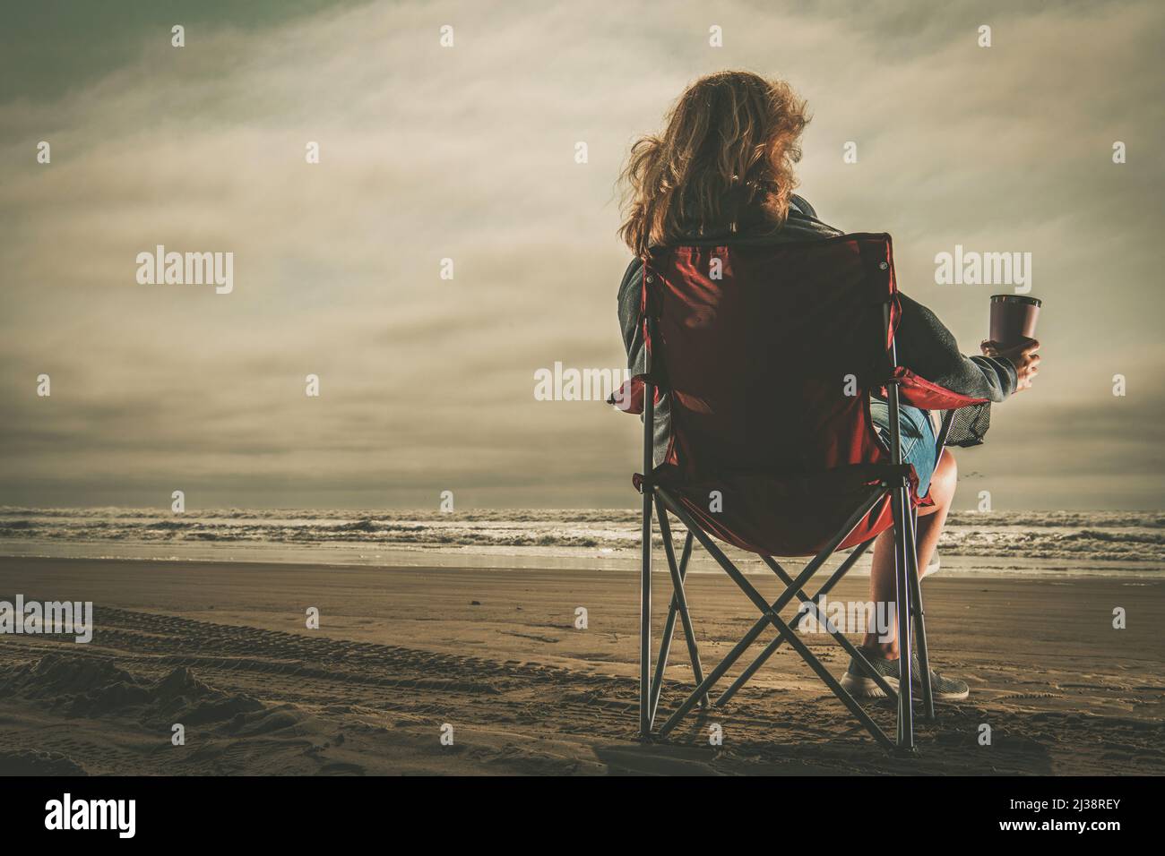 Kaukasische Frau mit Kaffee in der Hand, die Freizeit am Strand genießt. Sitzen auf einem Strandstuhl und den Sonnenuntergang beobachten. Stressfreies Leben. Stockfoto
