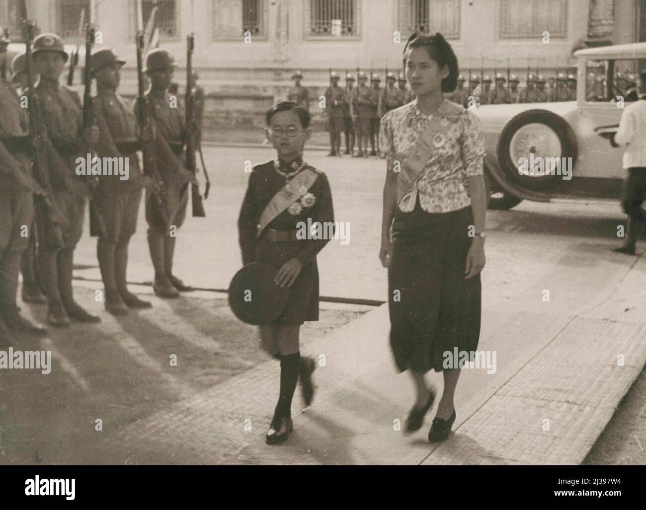 Der junge König von Siam präsentiert Farben -- der jüngere Bruder des Königs Bhumibol und seine Schwester kommen zur Parade. Der junge König Ananda von Siam erhält eine allmähliche Einführung in die vielen verantwortungsvollen Aufgaben, die er übernehmen muss, wenn er älter wird. Obwohl er erst dreizehn Jahre alt ist, nimmt er bereits jetzt eine Hauptaktion in einer Reihe von öffentlichen Zeremonieneien ein. Vor kurzem war er zum Beispiel bei einem großen Jugendlicher dabei. Parade in Bangkok, wo er den Einheiten Yuvajon und Yuvanari Farben präsentierte. 23. Januar 1939. (Foto von Associated Press Photo). Stockfoto
