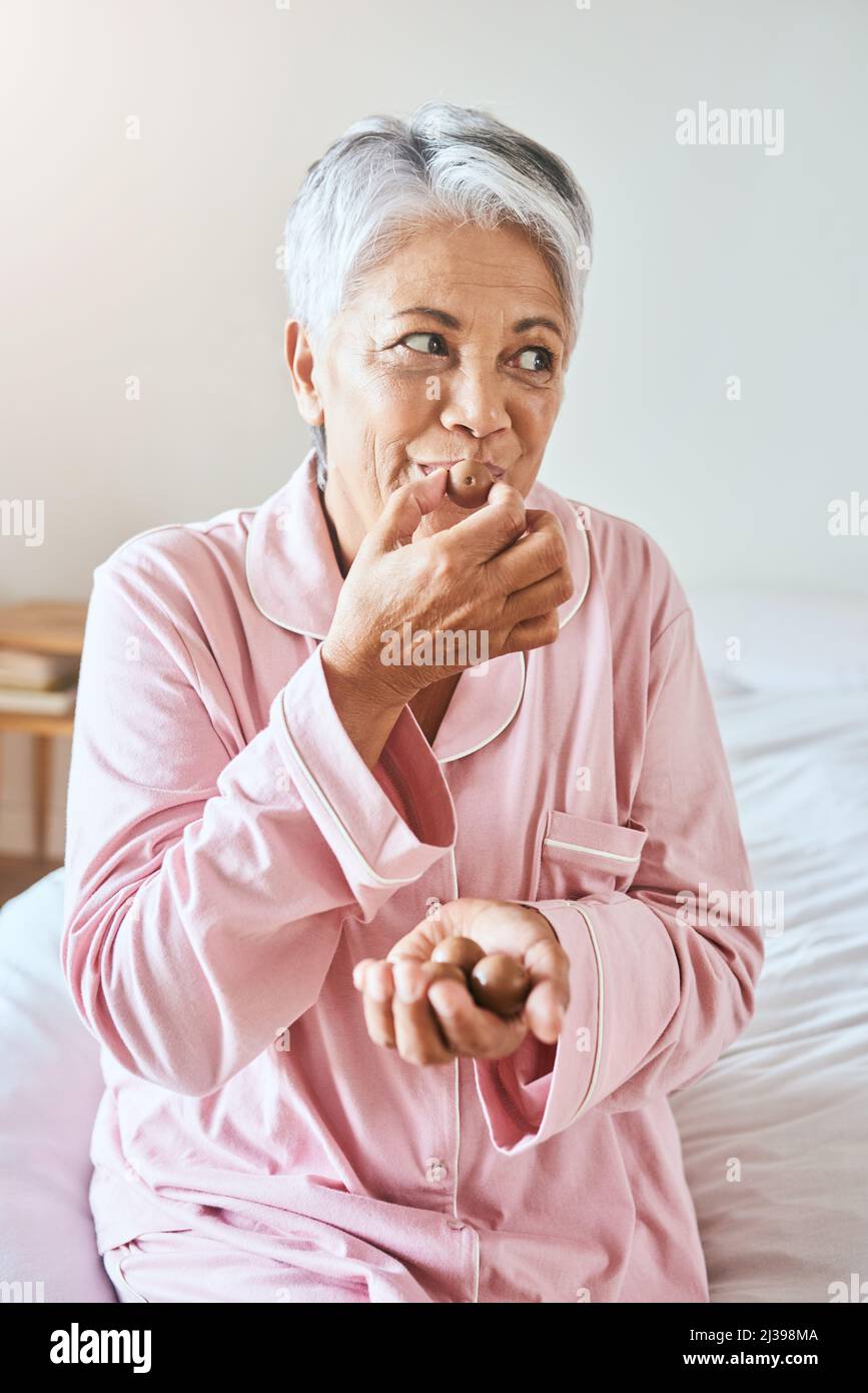 Oh ja, ich habe definitiv einen süßen Zahn. Aufnahme einer fröhlichen reifen Frau, die ein Stück Schokolade genießt, während sie zu Hause auf einem Bett sitzt Stockfoto