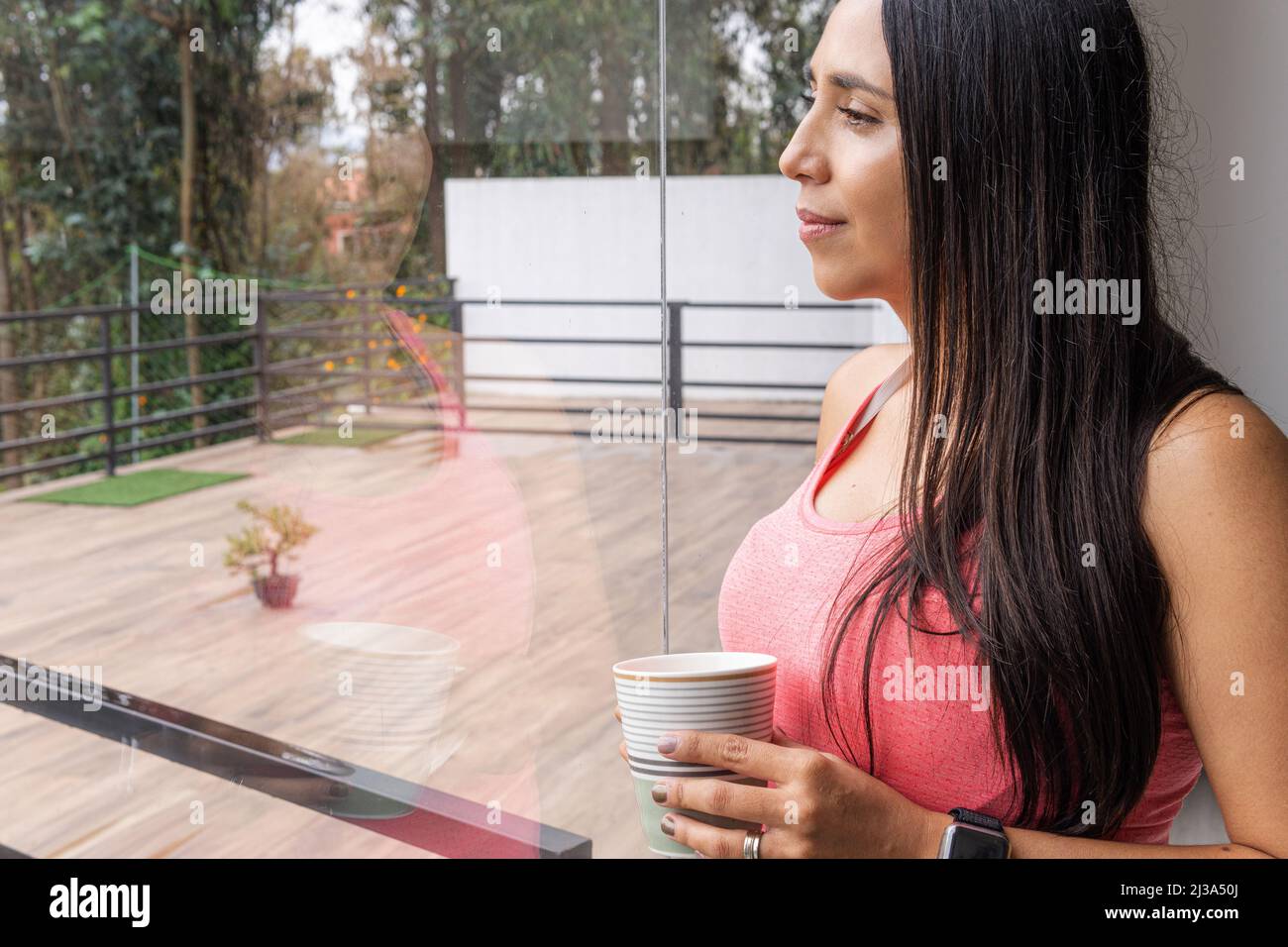 Die junge lateinische Frau trinkt eine Tasse Kaffee oder Tee und blickt durch ein Fenster auf die Terrasse, wobei sich ihr Bild im Glas widerspiegelt Stockfoto