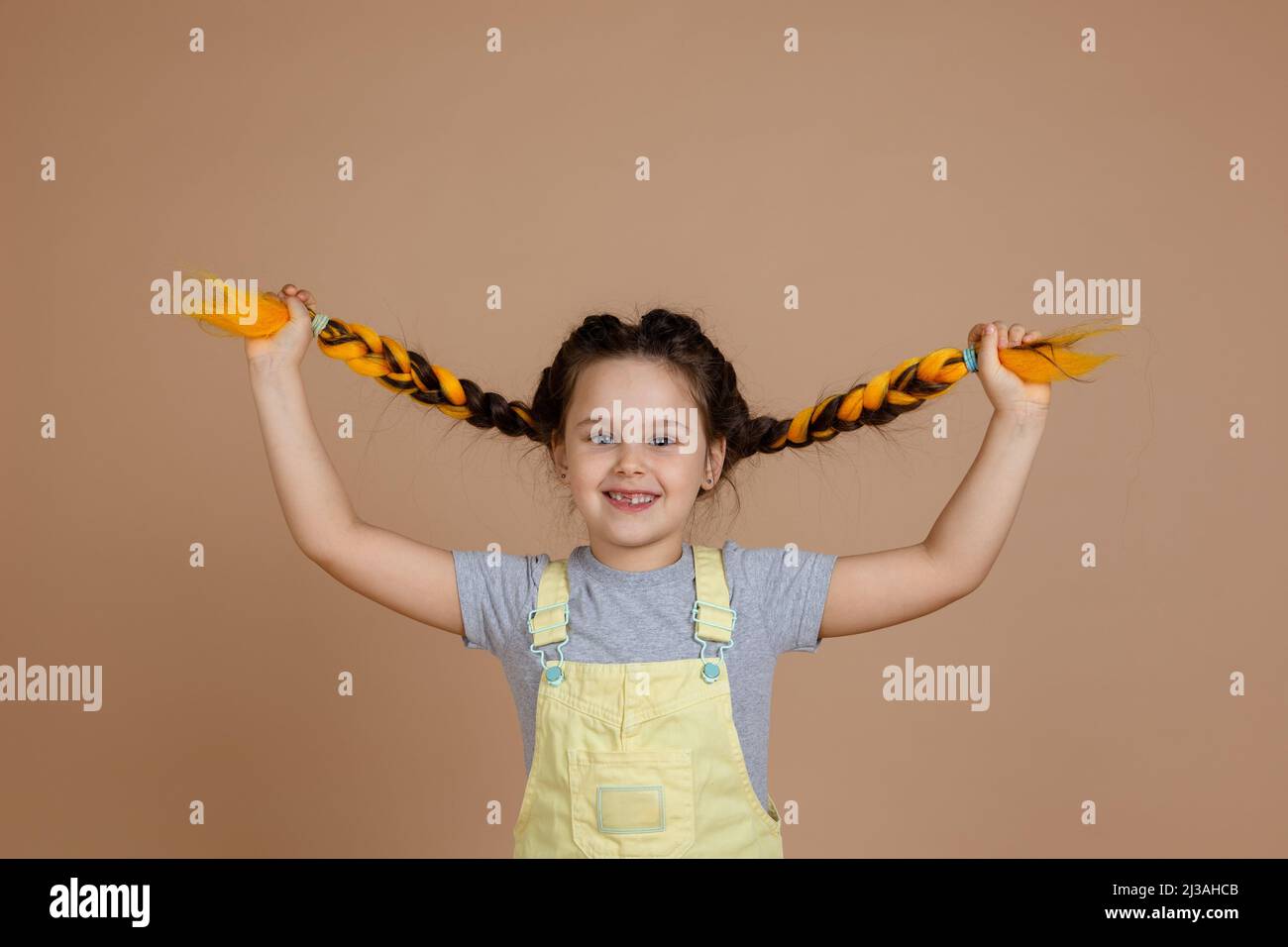 Zufriedenes glückseliges kleines Kind mit Kanekalon-Zöpfen von gelber Farbe, ziehen Zöpfe mit Händen Blick auf Kamera lächelnd tragen gelben Jumpsuit Stockfoto