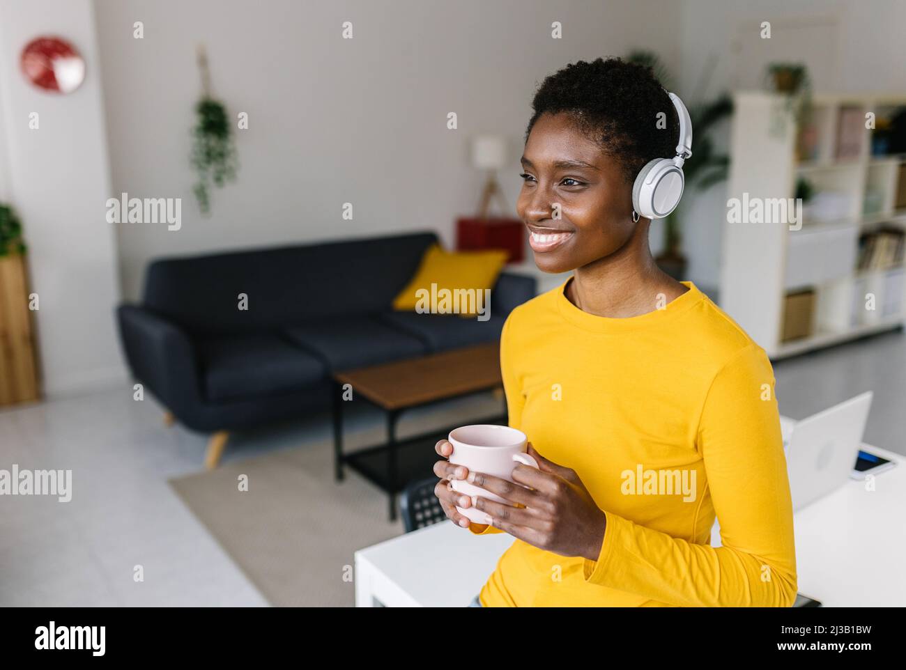 Multirassische Frau, die zu Hause Kaffee trinkt und Musik hört, mit Kopfhörern Stockfoto