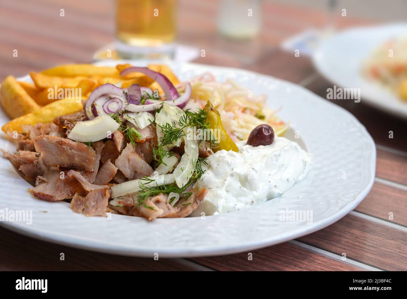 Griechisches Gyros-Gericht aus gebratenem, in Scheiben geschnittenem Fleisch mit Pommes frites, Zwiebeln, Krautsalat und Tsatziki auf einem weißen Teller in einem Restaurant im Freien, Nahaufnahme, s. Stockfoto