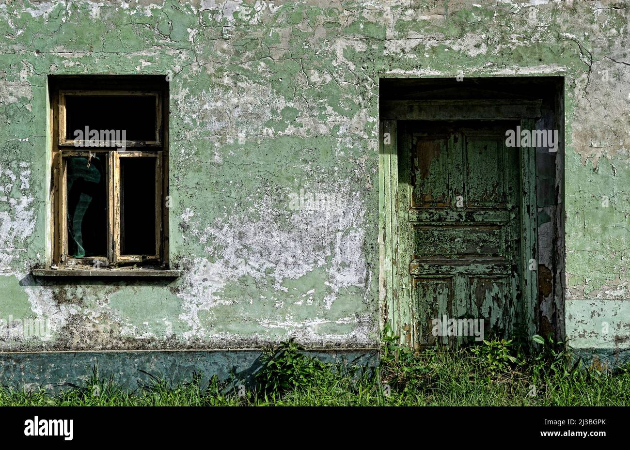 Eine alte Holztür und ein kaputtes Fenster mit einem verfallenen antiken dekorativen weißen Holzrahmen, die an der Wand des Familienhauses angebracht sind. Stockfoto