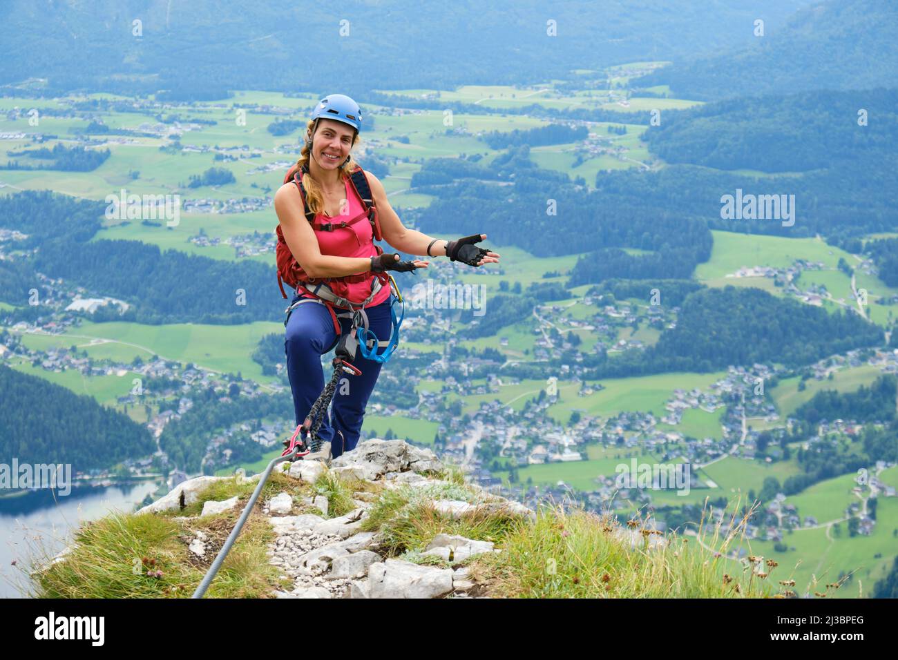 Frau mit Helm, Rucksack und Klettersteig-Ausrüstung, auf der Sisi Loser-Panorama in Österreich, lächelt und zeigt mit den Händen auf kleine Dörfer. Stockfoto