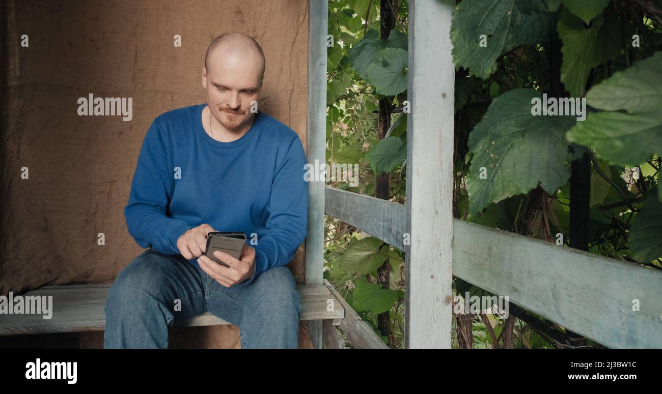 Der Mann kommuniziert online mit seinem Telefon, während er auf der Veranda des Dorfhauses sitzt Stockfoto