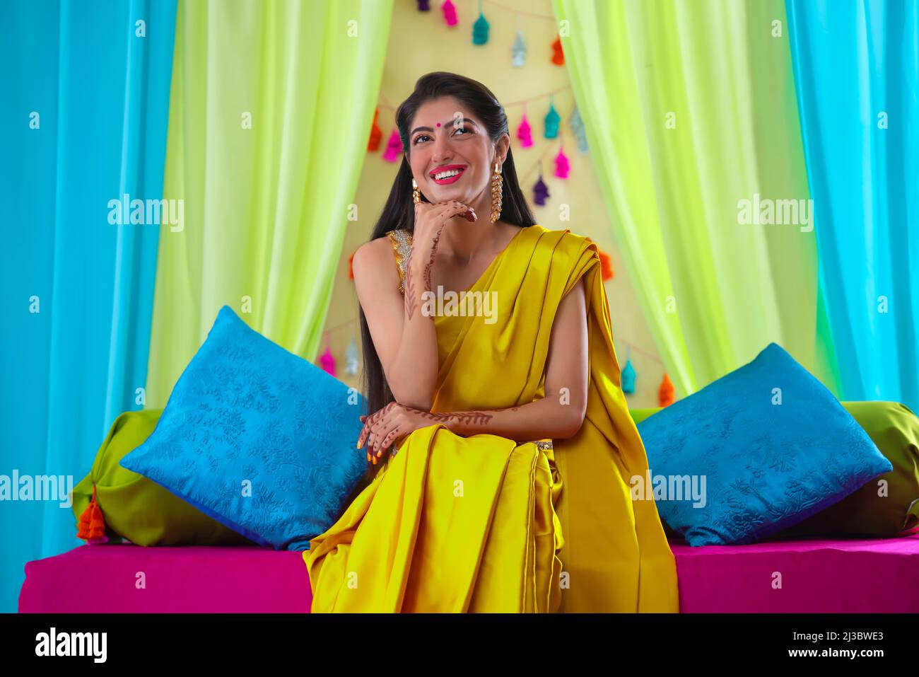 Indische Braut in gelbem Saree, die mit der Hand am Kinn auf der Bühne sitzt Stockfoto