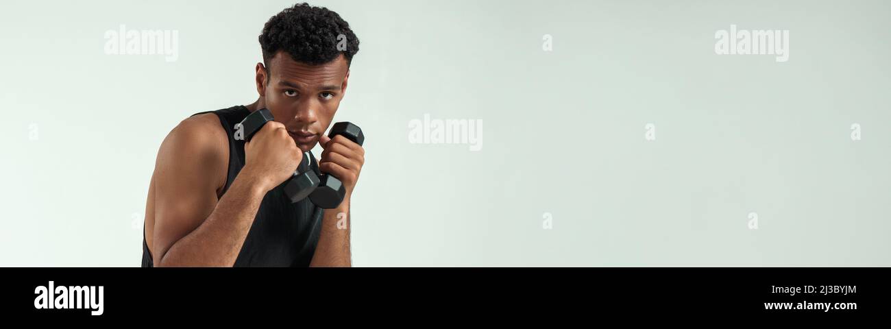 Perfekter Körper. Muskulöser junger afrikanischer Mann, der mit Hanteln trainiert, während er vor grauem Hintergrund steht Stockfoto