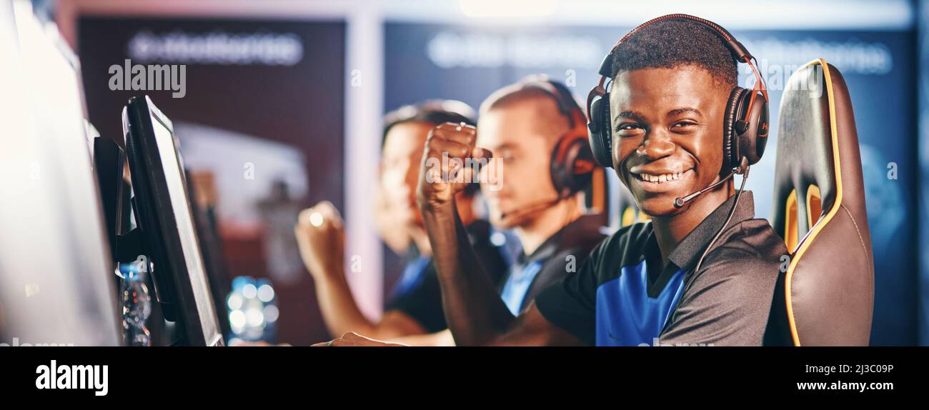 Seitenansicht eines fröhlichen afrikanischen Mannes, professioneller Cybersport-Gamer, der Kopfhörer trägt und die Kamera anschaut und lächelt, während er mit dem Team mitmacht Stockfoto