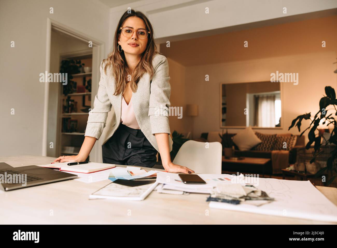 Professionelle Designerin, die in ihrem Heimbüro arbeitet. Junge Geschäftsfrau, die hinter ihrem kreativen Schreibtisch auf die Kamera schaut. Weibliche Designerin p Stockfoto