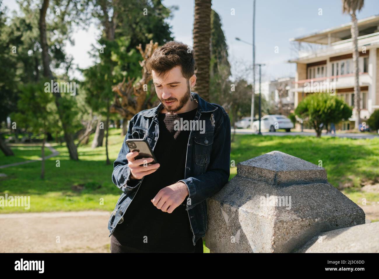 Junger Mann mit blauer jean-Jacke, der sich in einem Park an einen dekorativen Stein lehnt. Suchen nach Scrollen, Nachrichten oder Ansehen von Videos. Stockfoto