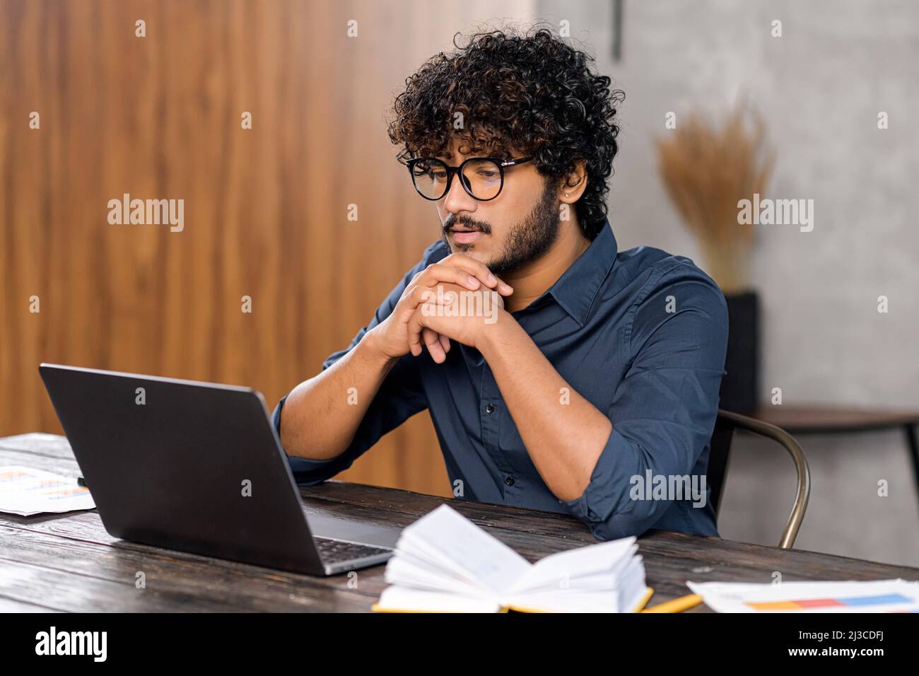 Fokussierter und besorgter indischer Mann in schickem Casual-Hemd, der auf den Laptop-Bildschirm schaut. Ernsthafter Typ, der am Schreibtisch sitzt und sich auf den Händen am Kinn ruht, während er Zweifel spürt und schwierige Aufgaben löst Stockfoto