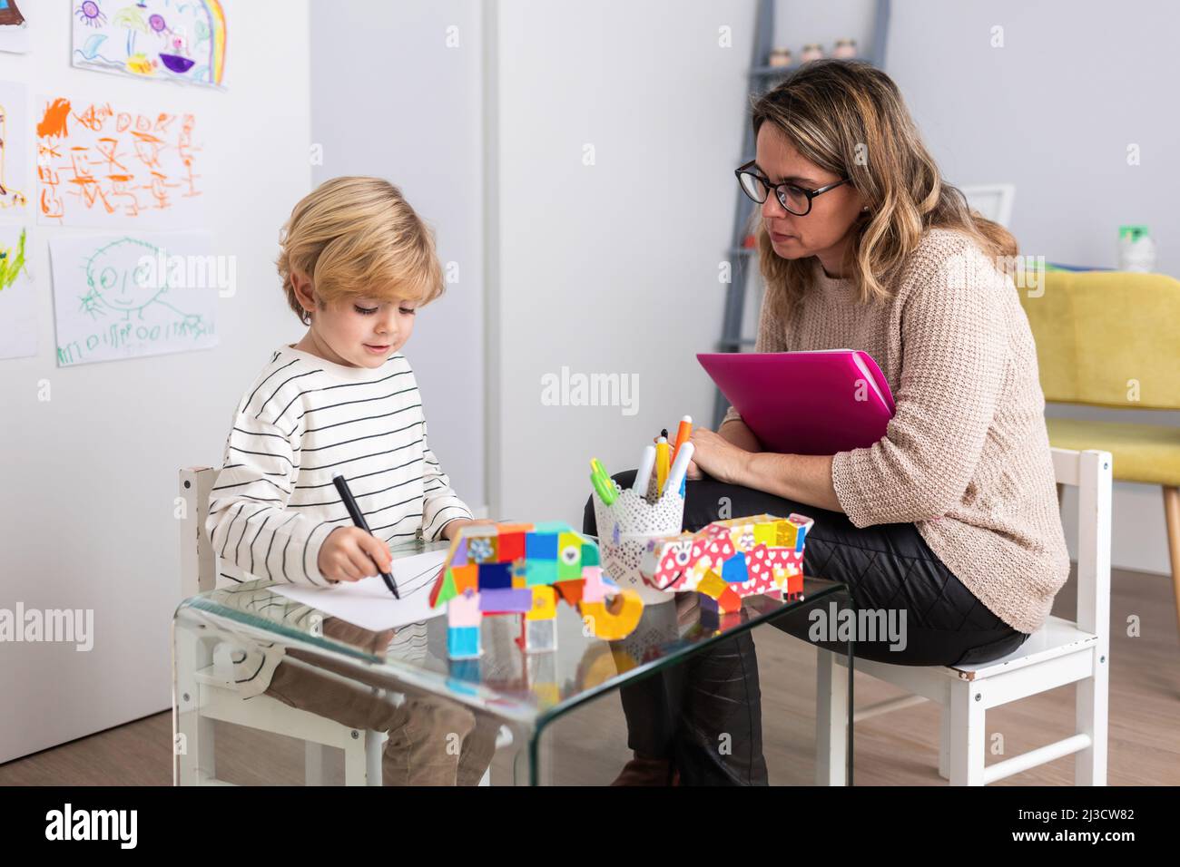 Fokussierte weibliche Psychologin mit Ordnern in der Nähe des Jungen sitzen Zeichnung auf Papier am Tisch mit Spielzeug während der Verabredung in hellen Raum Stockfoto