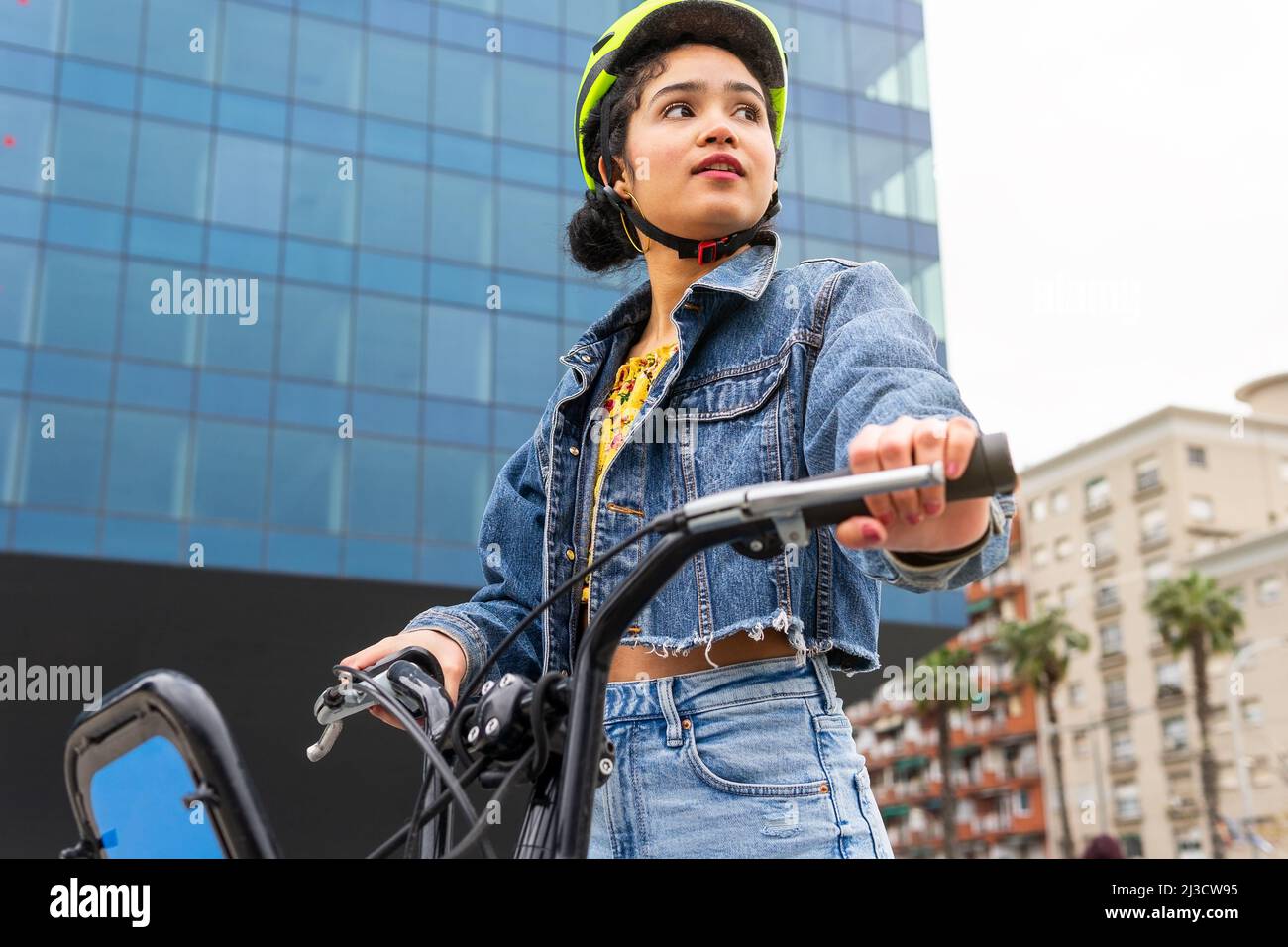 Von unten kolumbianische junge Dame trägt trendige Denim tragen und Schutzhelm stehen mit Fahrrad in der Nähe von modernen Glasgebäude während des aktiven Hobby p Stockfoto