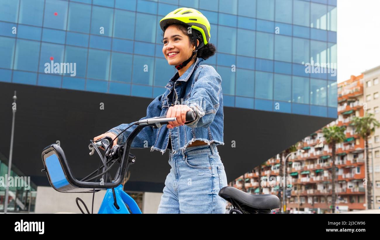Fröhliche kolumbianische junge Dame trägt trendige Denim-Kleidung und Schutzhelm stehen mit Fahrrad in der Nähe von modernen Glasgebäude während des aktiven Hobby pra Stockfoto
