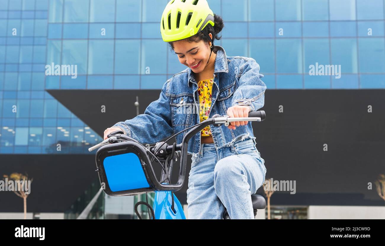 Fröhliche kolumbianische junge Dame in trendigen Denim-Kleidung und Schutzhelm, die während des aktiven Hobbypraktikums ein Fahrrad in der Nähe eines modernen Glasgebäudes fährt Stockfoto
