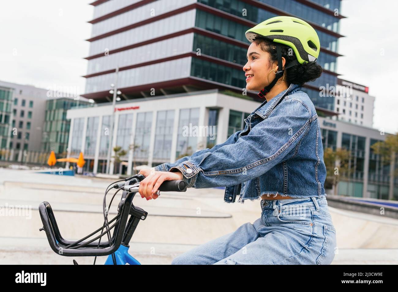 Fröhliche kolumbianische junge Dame in trendigen Denim-Kleidung und Schutzhelm, die während des aktiven Hobbypraktikums ein Fahrrad in der Nähe eines modernen Glasgebäudes fährt Stockfoto