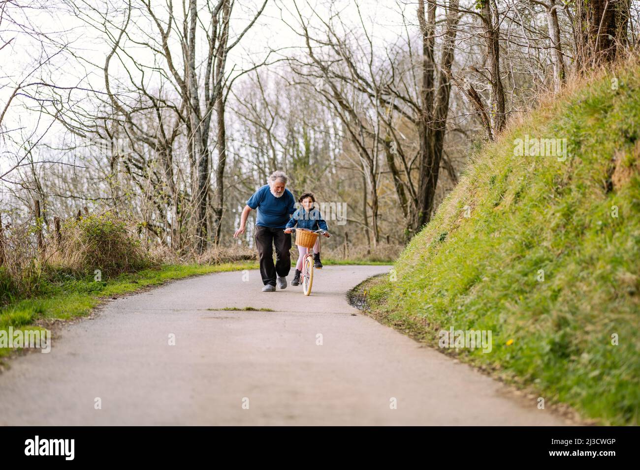 Entfernter fürsorglicher Großvater, der aktiven Enkelin hilft, Fahrrad mit Korb auf der Straße auf dem Land mit grünen Bäumen am Sommertag zu fahren Stockfoto