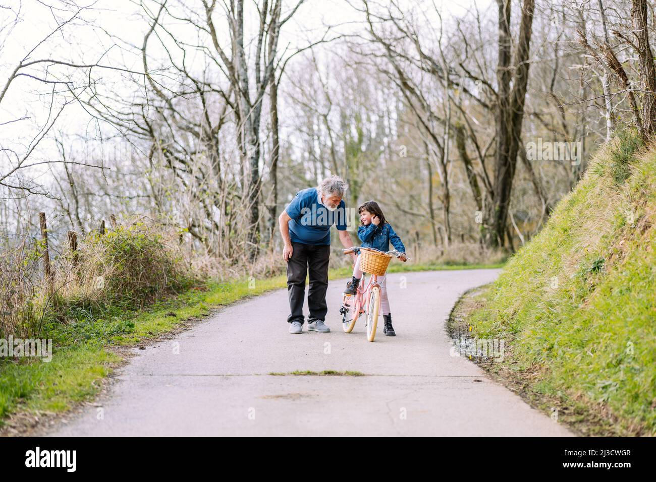 Entfernter fürsorglicher Großvater, der aktiven Enkelin hilft, Fahrrad mit Korb auf der Straße auf dem Land mit grünen Bäumen am Sommertag zu fahren Stockfoto