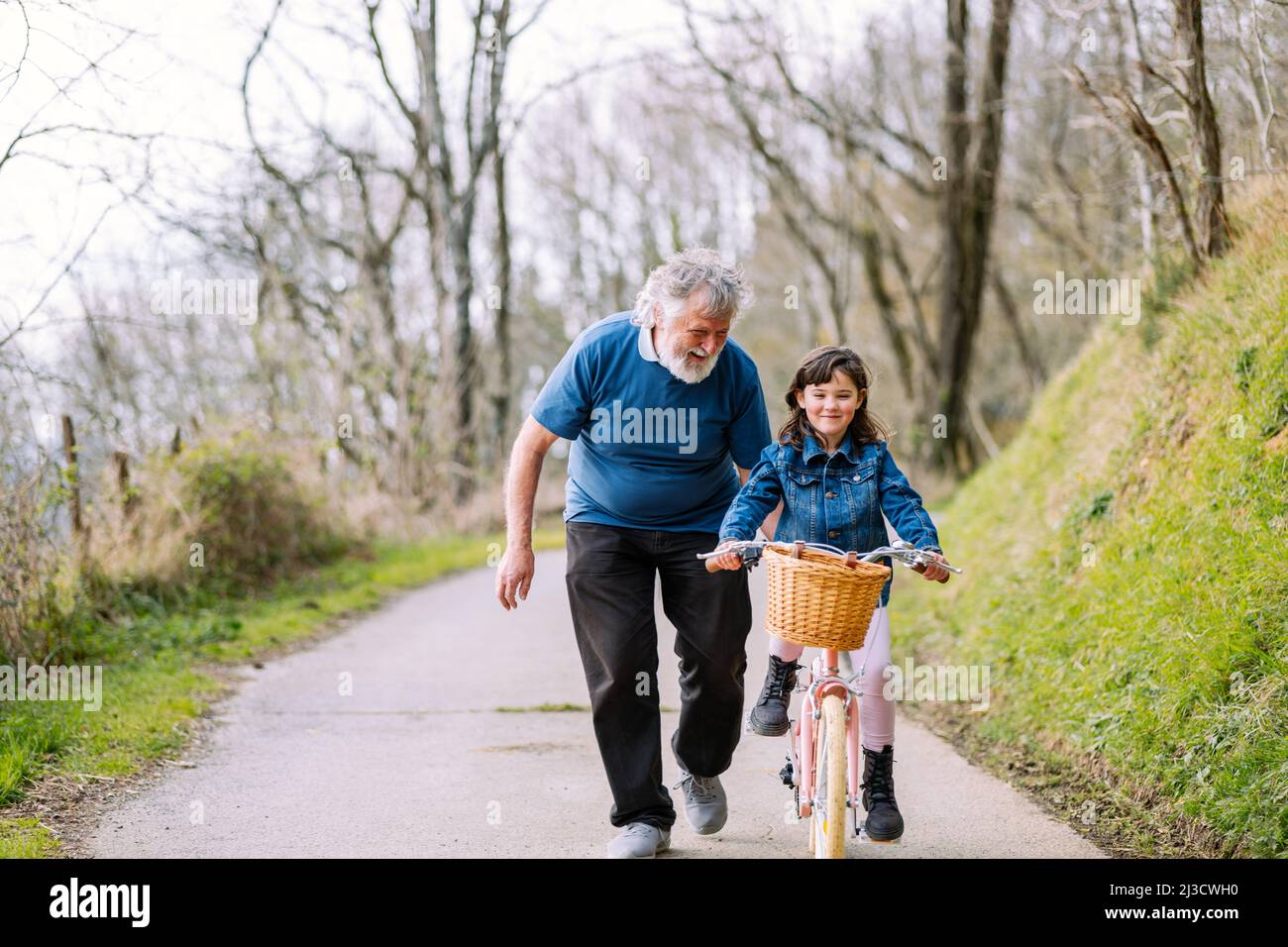 Entfernter fürsorglicher Großvater, der aktiven Enkelin hilft, Fahrrad mit Korb auf der Straße auf dem Land mit grünen Bäumen am Sommertag zu fahren Stockfoto