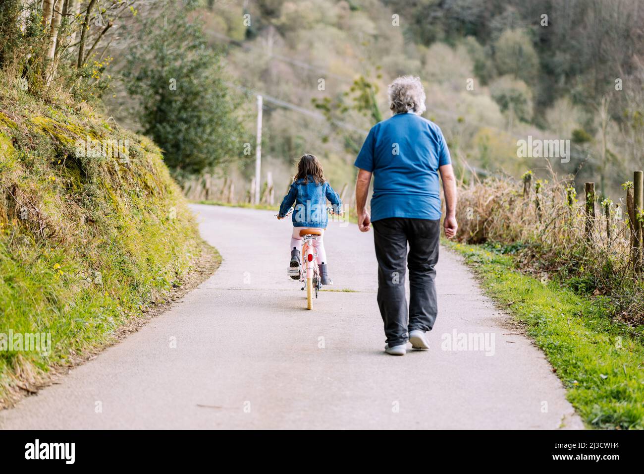 Rückansicht des unkenntlich fürsorglichen Großvaters, der aktiven Enkelin beim Fahrradfahren mit Korb auf der Straße auf dem Land mit grünen Bäumen auf dem Summ hilft Stockfoto