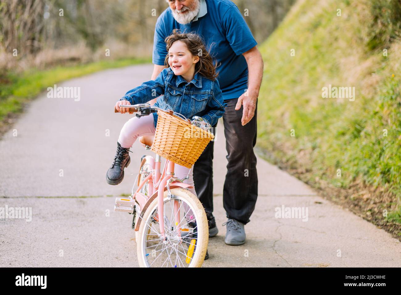 Fürsorglicher, zugeschnittenes Großvater, der aktiven Enkelin beim Fahrradfahren mit Korb auf der Straße auf dem Land mit grünen Bäumen am Sommertag hilft Stockfoto